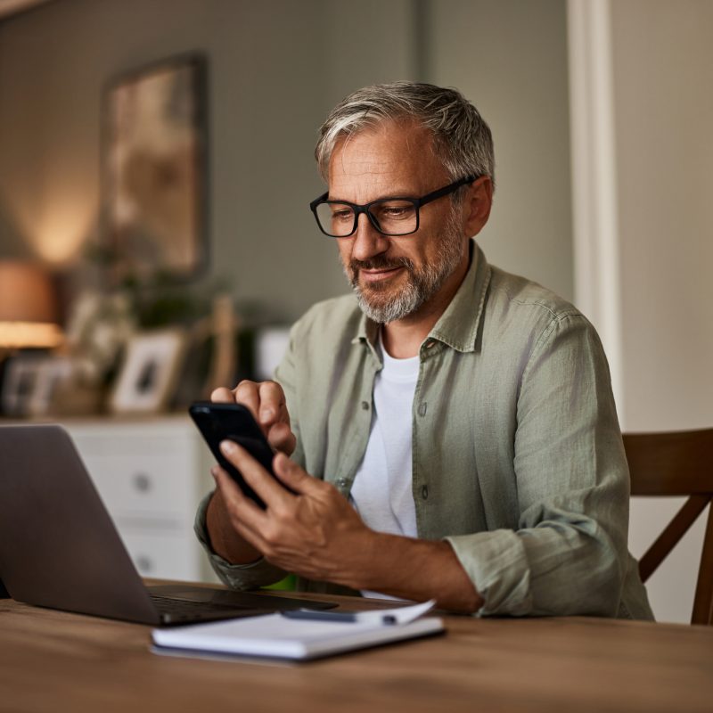 A man working from home over a laptop and using a mobile phone sitting at the wooden table.