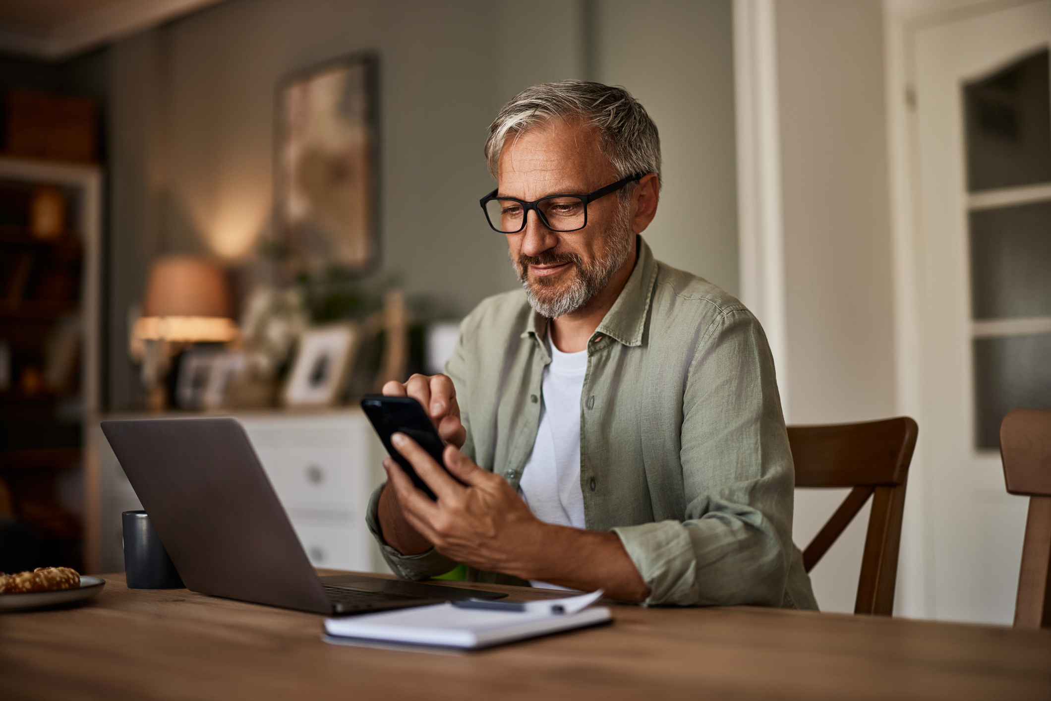 A man working from home over a laptop and using a mobile phone sitting at the wooden table.