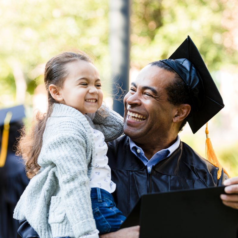 Image: male student at graduation holding his smiling young daughter in one arm and his diploma in the other.