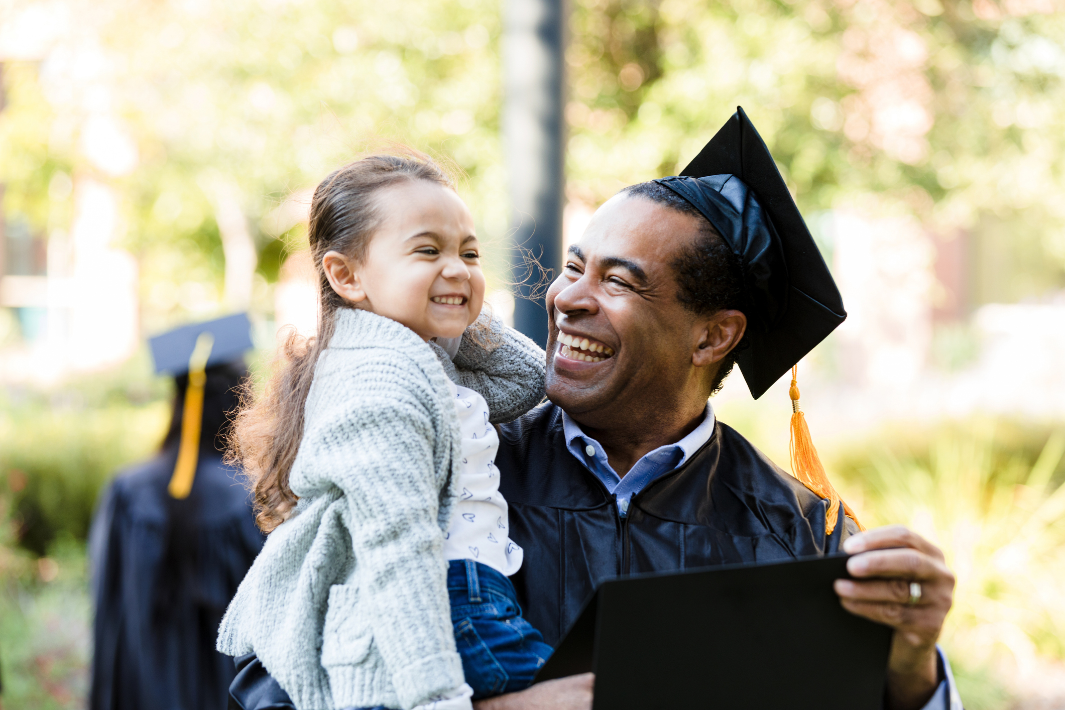 Image: male student at graduation holding his smiling young daughter in one arm and his diploma in the other.