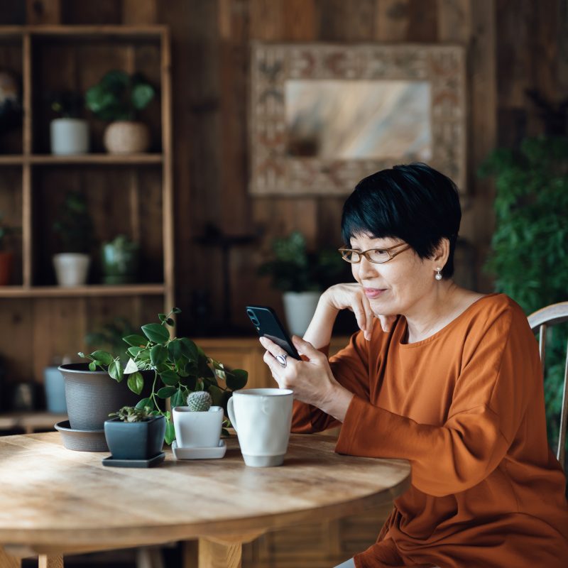 Image: Older Asian woman sitting at kitchen table with her mobile phone.